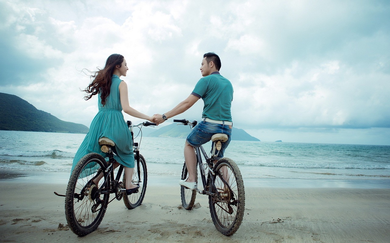photo of young couple riding bikes and holding hands on the beach